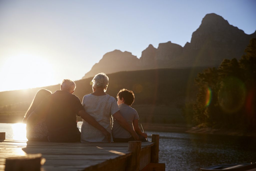 Grandchildren With Grandparents Sitting On Wooden Jetty By Lake